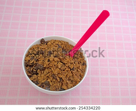 A bowl of organic whole grain raisin cereal with skim milk and a pink spoon inserted on a checkerboard table cloth.