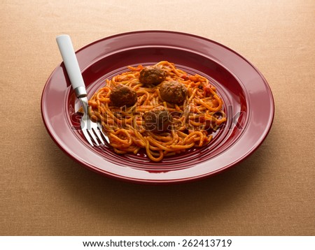 A spaghetti and meatball TV dinner on a red plate with a fork on a tan table cloth illuminated by window light.
