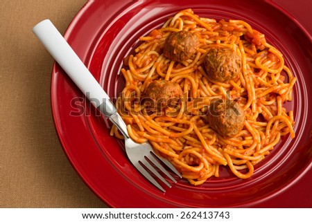 Top view of a spaghetti and meatball TV dinner on a red plate with a fork on a tan table cloth.