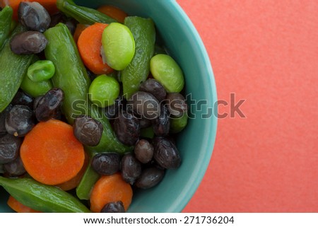 Close view of buttered carrots, black beans, edamame and snap peas in a bowl atop an orange background.