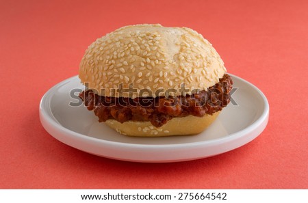 A freshly made sloppy joe sesame seed roll sandwich on a small plate atop an orange table top.