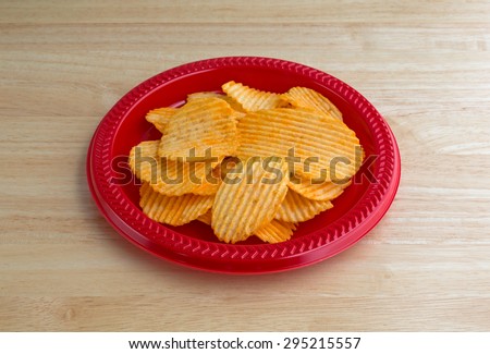 A red plastic plate with a serving of cheddar cheese flavored potato chips on a wood table top illuminated with natural light.