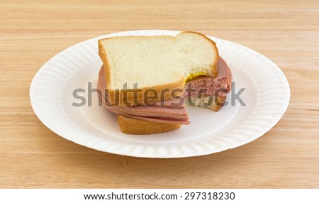 A several slices thick bitten bologna sandwich with mustard and white bread on a paper plate.