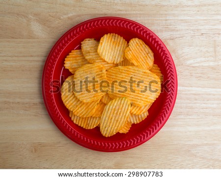 Top view of a red plastic plate with a serving of cheddar cheese flavored potato chips on a wood table top illuminated with natural light.
