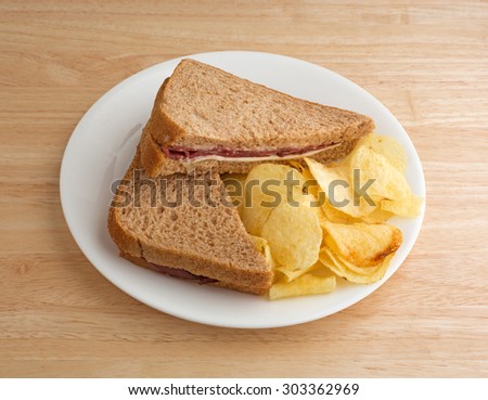 A roast beef wheat bread sandwich with white cheese and mayonnaise plus potato chips on a white plate atop a wood table top.