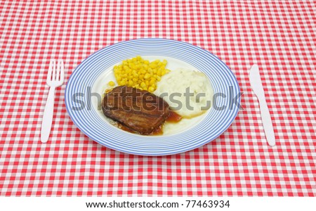 Salisbury steak meal with corn and potato on a blue striped plate with plastic silverware on a red checkerboard cloth.