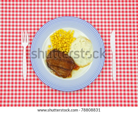 Salisbury steak dinner with corn and potato on a blue striped plate with plastic silverware on a red checkerboard cloth.