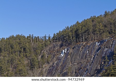 A steep rock face side of mountain surrounded by forest with ice.