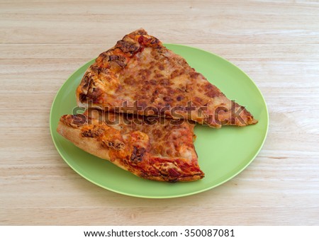 Two leftover cold cheese pizza slices on a green plastic plate atop a wood table top.