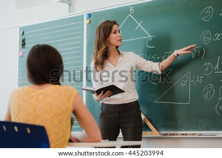 Teacher or docent or educator giving while lesson in front of a blackboard or board a sheet of paper and educate or teaching students or pupils or mates in a school or class