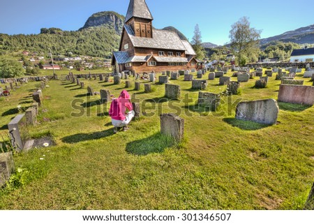 Woman in red hooded jacket kneels praying  at graveside in cemetery of small wooden church with tower