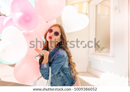 Charming long-haired girl in stylish sunglasses sending kiss while posing with heart balloons. Adorable young woman with curly hair and red lipstick wearing denim jacket, having fun at the party