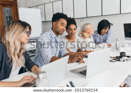 Long-haired curly student typing on keyboard, sitting beside african friend in university. Indoor portrait of it-specialists working together in light office and smiling.