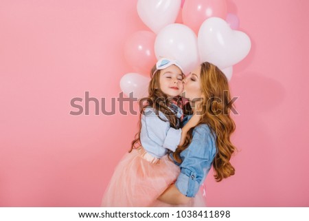 Long-haired mother and daughter in same denim shirts cute embrace at festive event with white balloons. Portrait of young beautiful woman kissing her child at birthday party on pink background