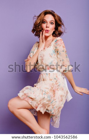 Barefooted graceful girl dancing on purple background with amazed face expression. Lovely young lady in dress with flower pattern posing emotionally in studio.