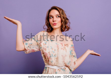 Glamorous young female model looking away with smile, waving hands on purple background. Stunning curly girl with short haircut wears dress with flower pattern.