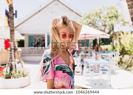 Romantic girl in pink attire touching her blonde hair and smiling on blur background in sunny day. Tanned european lady in round glasses expressing happiness during rest at summer resort.