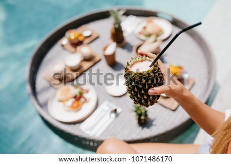 Tanned woman holding sweet pineapple cocktail. Female model posing during lunch in pool.