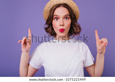 Close-up portrait of surprised dark-eyed girl in summer hat. Indoor shot of funny curly female model in white t-shirt posing with fingers up on purple background.