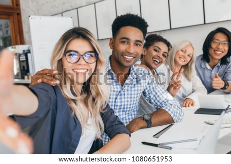 Black office worker in checkered shirt embracing blonde secretary girl while she making selfie. Young managers of international company having fun during meeting.