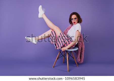 Good-looking caucasian girl with bright makeup sitting on chair. Relaxed female model in white shoes posing on purple background and waving legs.