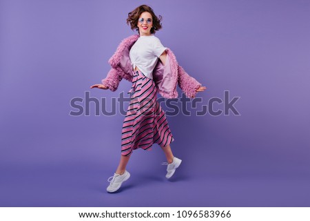 Laughing fascinating girl in white sneakers jumping on purple background. Full-length photo of enthusiastic young woman with wavy hair dancing in studio.
