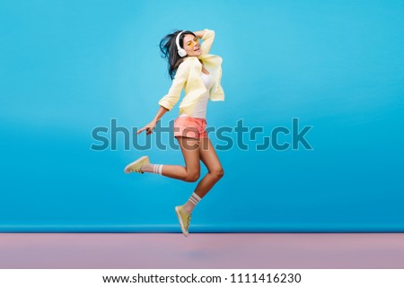 Full-length portrait of carefree brunette woman in shorts jumping while listening music. Indoor photo of adorable asian female model in yellow jacket fooling around in studio with blue walls.