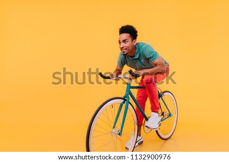 Trendy african man in white sneakers posing emotionally on bike. Indoor portrait of enthusiastic sportive guy sitting on bicycle.