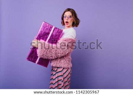 Adorable white young woman posing on purple background with sparkle gift box. Birthday girl with surprised face expression holding her present.