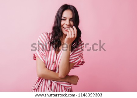 Excited brunette woman wears ring smiling on pink background. Indoor photo of gorgeous dark-haired girl in elegant striped dress.