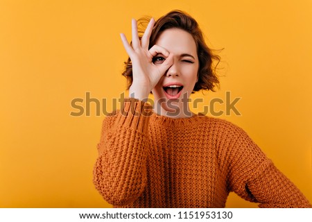 Blissful young woman with pale skin making funny faces during photosoot. Studio shot of pretty curly girl fooling around on bright orange background.