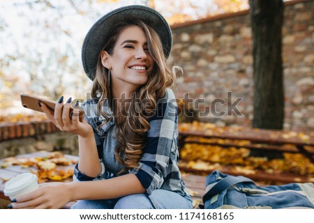 Pretty laughing girl with smartphone has a good time in autumn weekend. Outdoor portrait of lovable trendy lady with brown hair wears hat in october day.