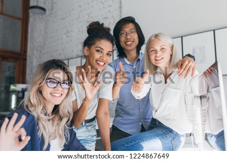 Asian male student spending time with university mates and posing in room with loft interior. Elegant female office worker in white shirt sits on table and having fun with co-workers.
