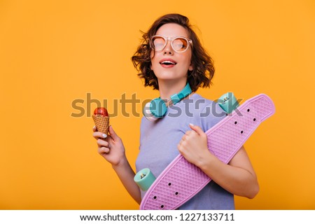 Interested dark-haired girl with blue earphones eating ice cream. Indoor photo of funny curly woman with skateboard isolated on yellow background.