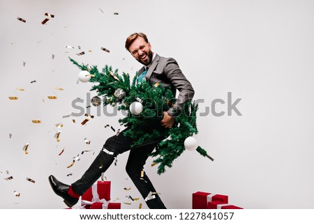 Blissful dark-haired man fooling around while preparing for new year. Laughing male model holding green christmas tree.