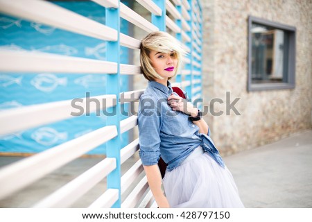 Portrait of a dreamy blond girl with short hair, bright pink lips and blue eyes wearing blue denim shirt,grey tulle skirt and marsala backpack leaning on blue and white stripes fence on the background