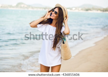 Cute stylish girl standing on a beach talking on a smartphone. Girl wears straw hat, beach straw bag and short white dress with open shoulders. She has brown sunglasses on. Ocean behind her.