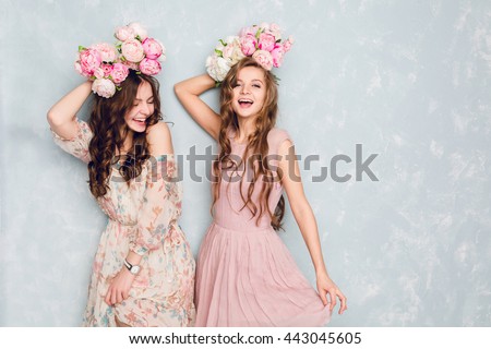 Two beautiful girls stand in a studio, play silly and have circlets of flowers on their heads. They wear light silk dresses. One is blond, and one is brunette. Both have curly hair.
