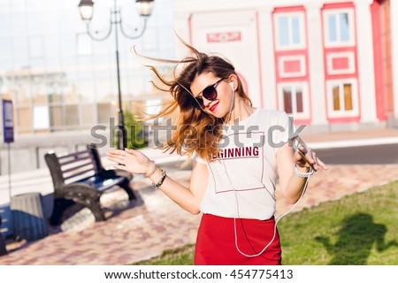 Horizontal portrait of a standing widely smiling and dancing young girl holding a smartphone and listening to music. Girl wears a white t-shirt, red skirt and dark sunglasses.