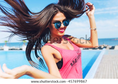Close-up portrait of attractive brunette girl with long hair  jumping to the camera near pool. She wears pink T-shirt with shorts and sunglasses. She looks very happy.