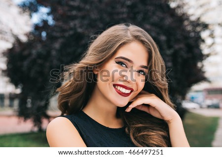 Closeup portrait of pretty girl with long curly hair on street on black trees background. She wears black dress, red lips. She is touching face and smiling to camera.