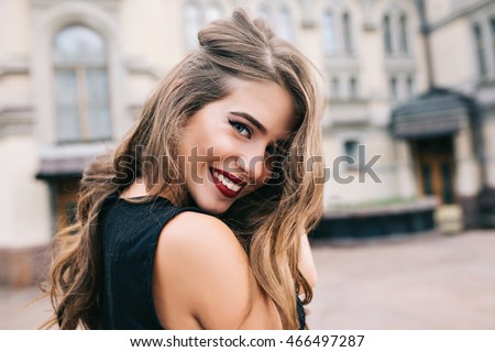 Closeup portrait of pretty girl with long curly hair smiling to camera in city on old building background. She wears black dress, red lips. View from back.