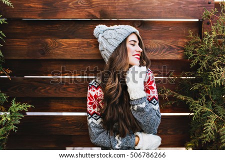 Portrait beautiful girl with long hair and red lips in warm winter clothes on wooden background. She is smiling to side and keeps eyes closed