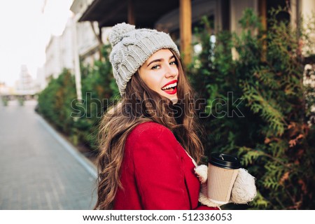 Portrait pretty girl with long hair in red coat walking on street with coffee to go. She looks excited to camera
