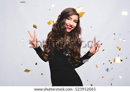 Young stylish woman on white background drinking champagne, celebrating new year, wearing black dress and yellow crown, happy carnival disco party, sparkling confetti, holding glass, having fun