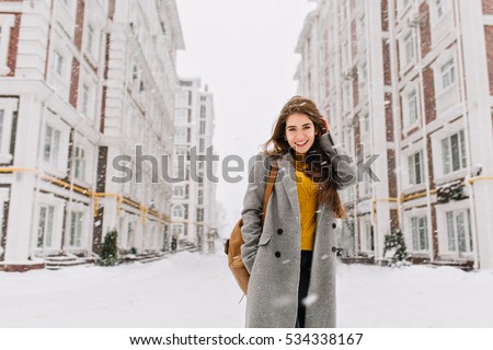 Charming young woman in coat with long brunette hair enjoying snowfall in big city. Cheerful emotions, smiling, christmas mood, positive face emotions, winter weather. Place for text