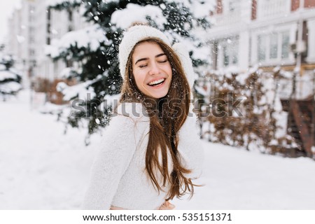 Happy winter moments of joyful young woman with long brunette hair, white winter clothes having fun on street in snowing time. Expressing positivity, true brightful emotions, smiling with closed eyes