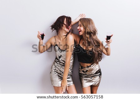 Two attractive girls, cheerful best friends dancing, having fun and drinking red wine on party. Wearing shiny dresses with paillettes, fashionable looking with beautiful wavy hair. Isolated.