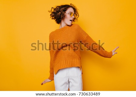 Gorgeous slim woman in white pants jumping on yellow background and looking away. Interested curly girl in oversize sweater having fun in studio.