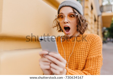 Shocked girl with brown eyes looking at phone screen with mouth open. Outdoor portrait of surprised young woman with curly hair wearing yellow knitted attire and holding smartphone.